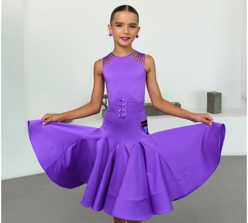 Young dancer in a vibrant purple ballroom dress with flared skirt and decorative straps, posing with arms extended to display the fullness of the outfit
