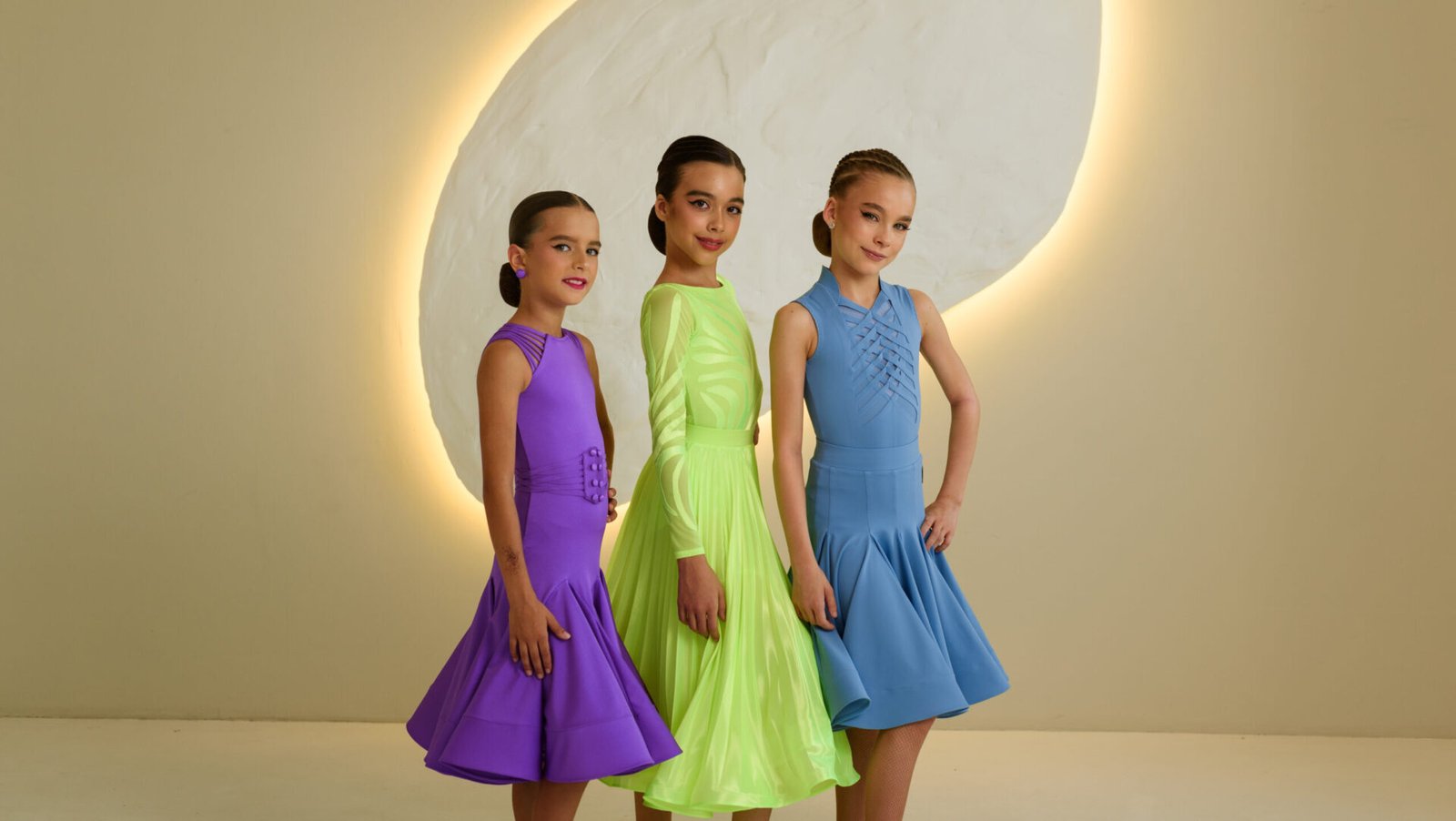 Three children wearing vibrant couture dresses in purple, neon green, and blue, standing in front of a beige wall with circular lighting. Each outfit features unique design elements and elegant styling.