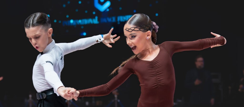 Young ballroom dance couple performing on stage, girl wearing a flowing maroon dress and boy in classic black‑and‑white attire during a competition.